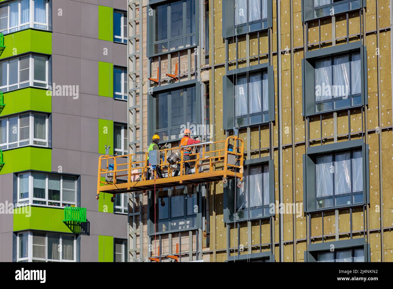 Male builders work on the facade of the building in a suspended cradle. Construction works. Stock Photo