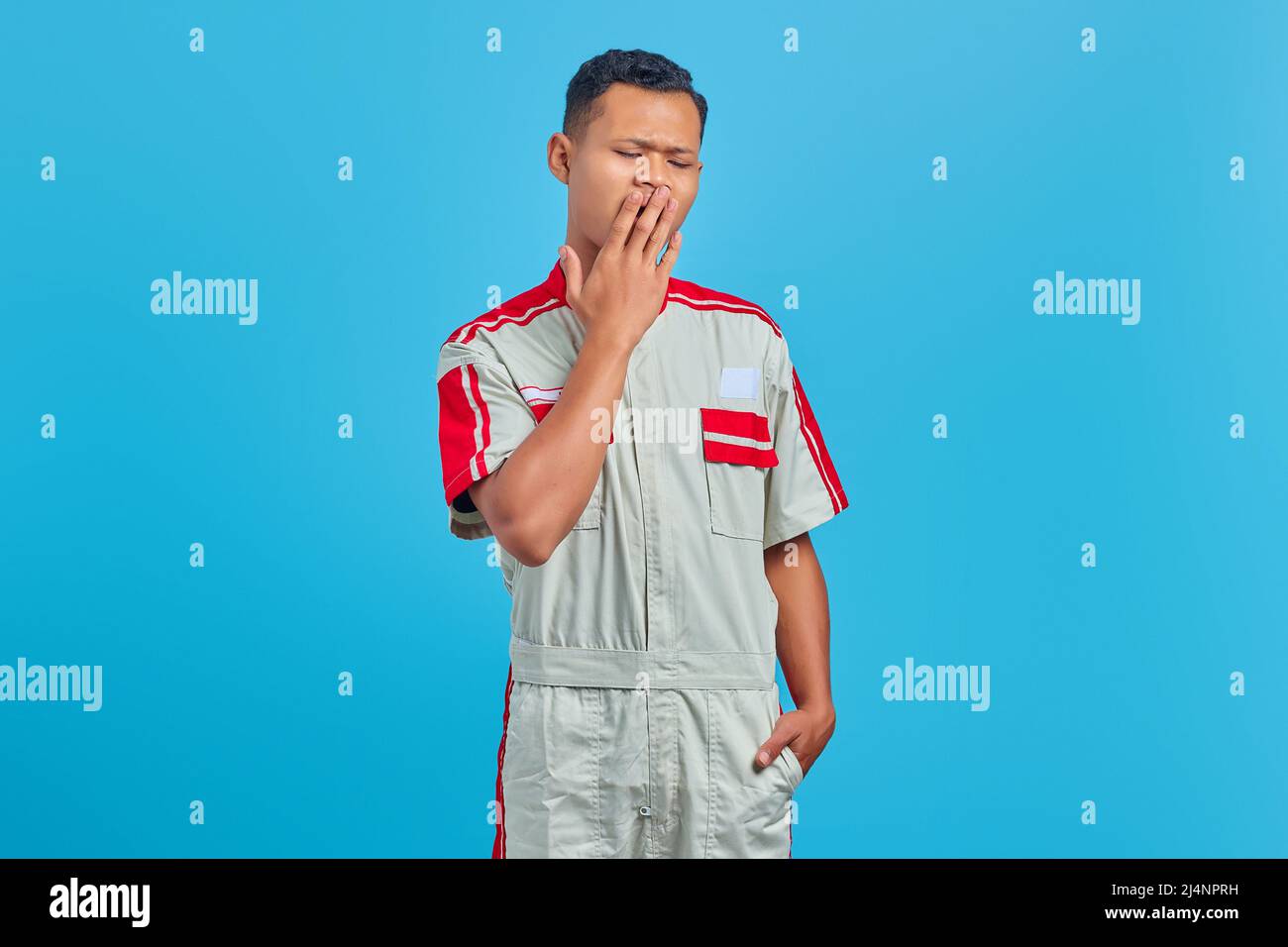 Portrait of young mechanic feeling sleepy and covering mouth with hand on blue background Stock Photo