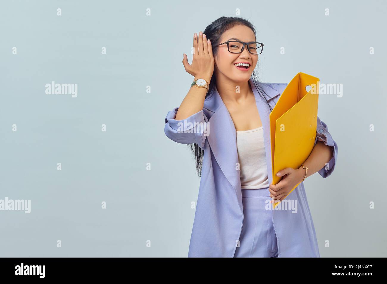 Portrait of serious young Asian business woman keeps hand near ear tries to overhear private conversation listens gossips while holding yellow folder Stock Photo