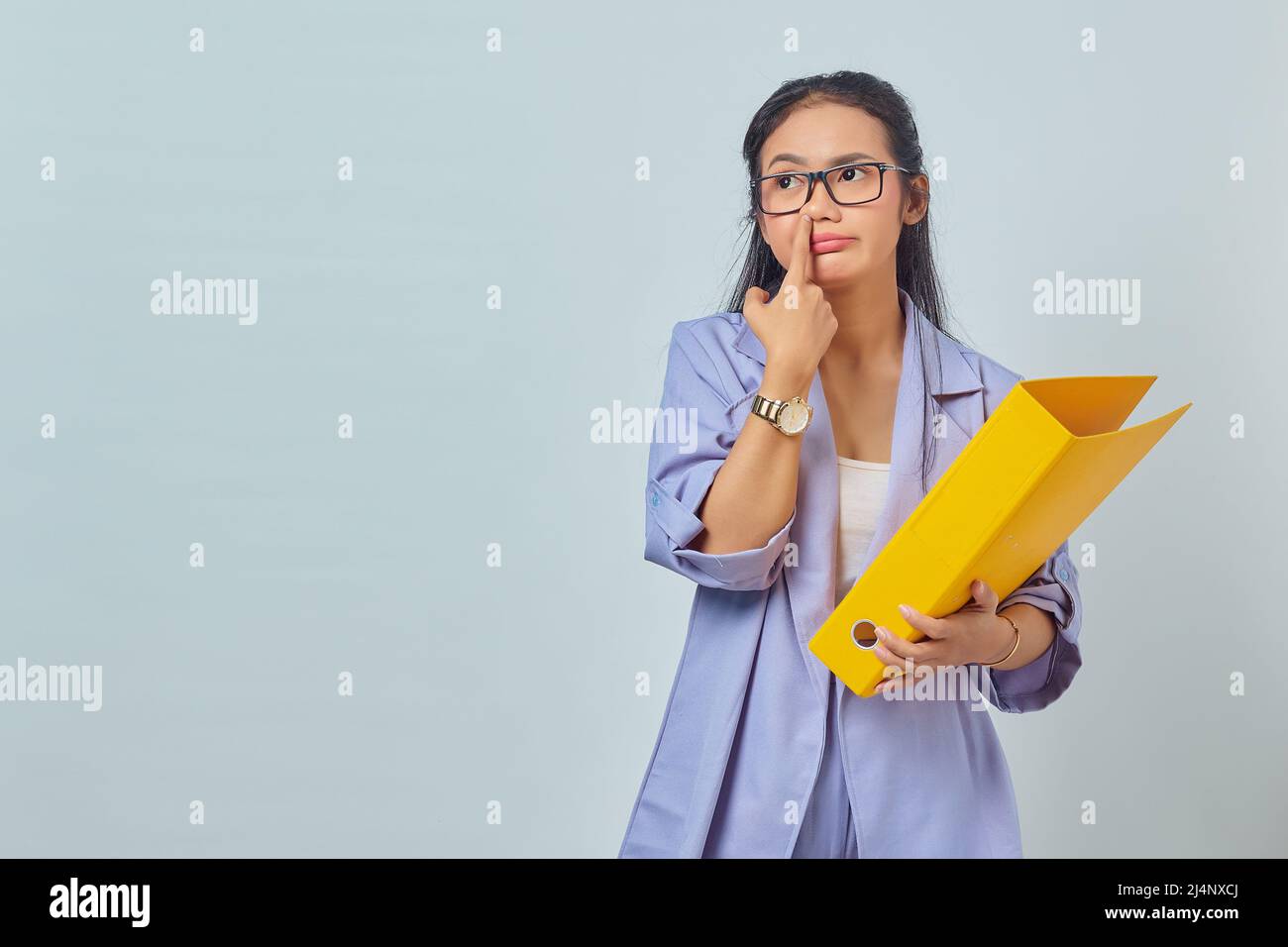 Portrait of funny young Asian woman thinking about something while carrying yellow folder and  picking her nose isolated on yellow background Stock Photo