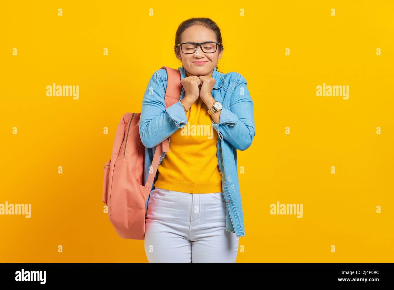 Excited young Asian student in denim clothes with backpack keeps hands under chin, remembers pleasant moment isolated on yellow background. Education Stock Photo