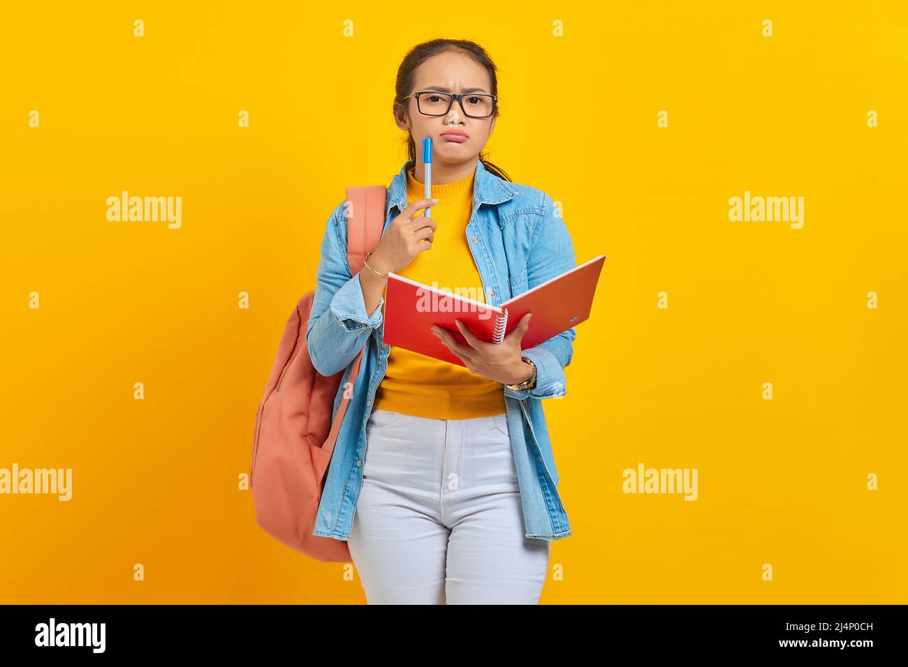 Beautiful young Asian woman student in casual clothes with backpack holding book and looks serious thinking about  creative idea isolated on yellow ba Stock Photo