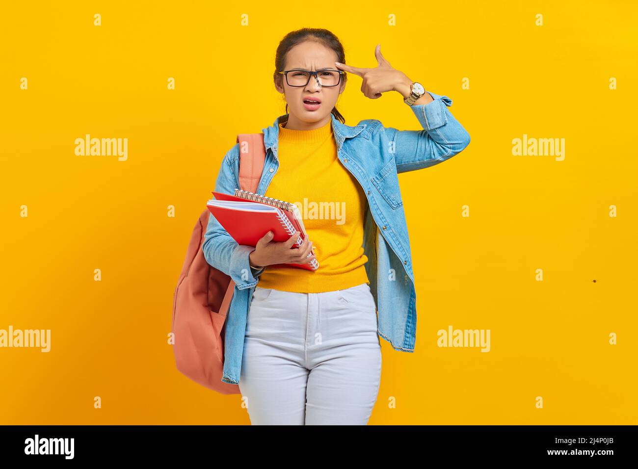 Beautiful young woman student in denim clothes with backpack,holding note book and Point fingers to head as if she about shoot herself isolated on yel Stock Photo