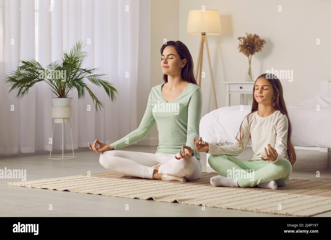 Beautiful woman and her little daughter are smiling while doing yoga together at home. Stock Photo