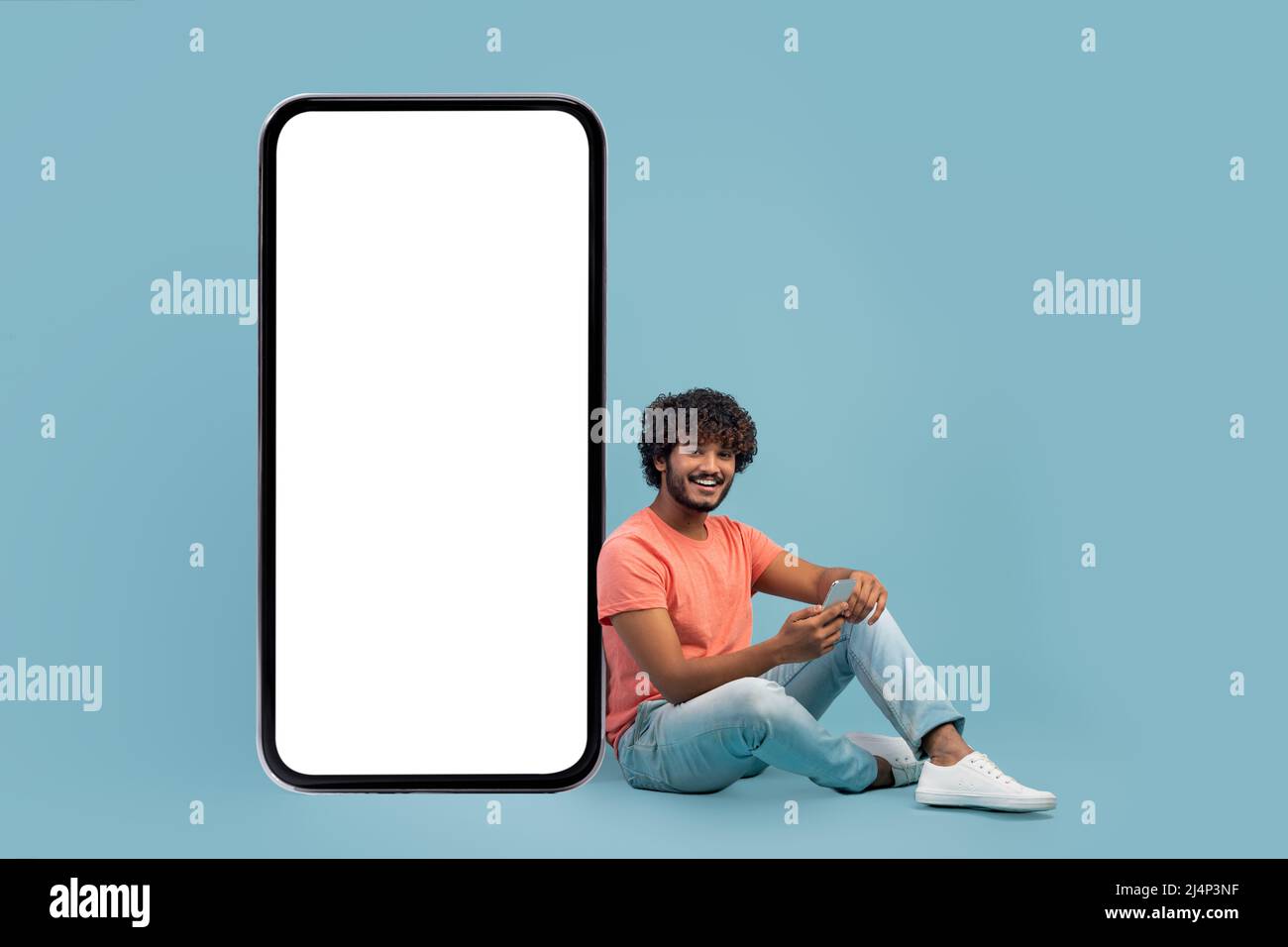 Handsome indian guy sitting by huge smartphone, using cell phone Stock Photo