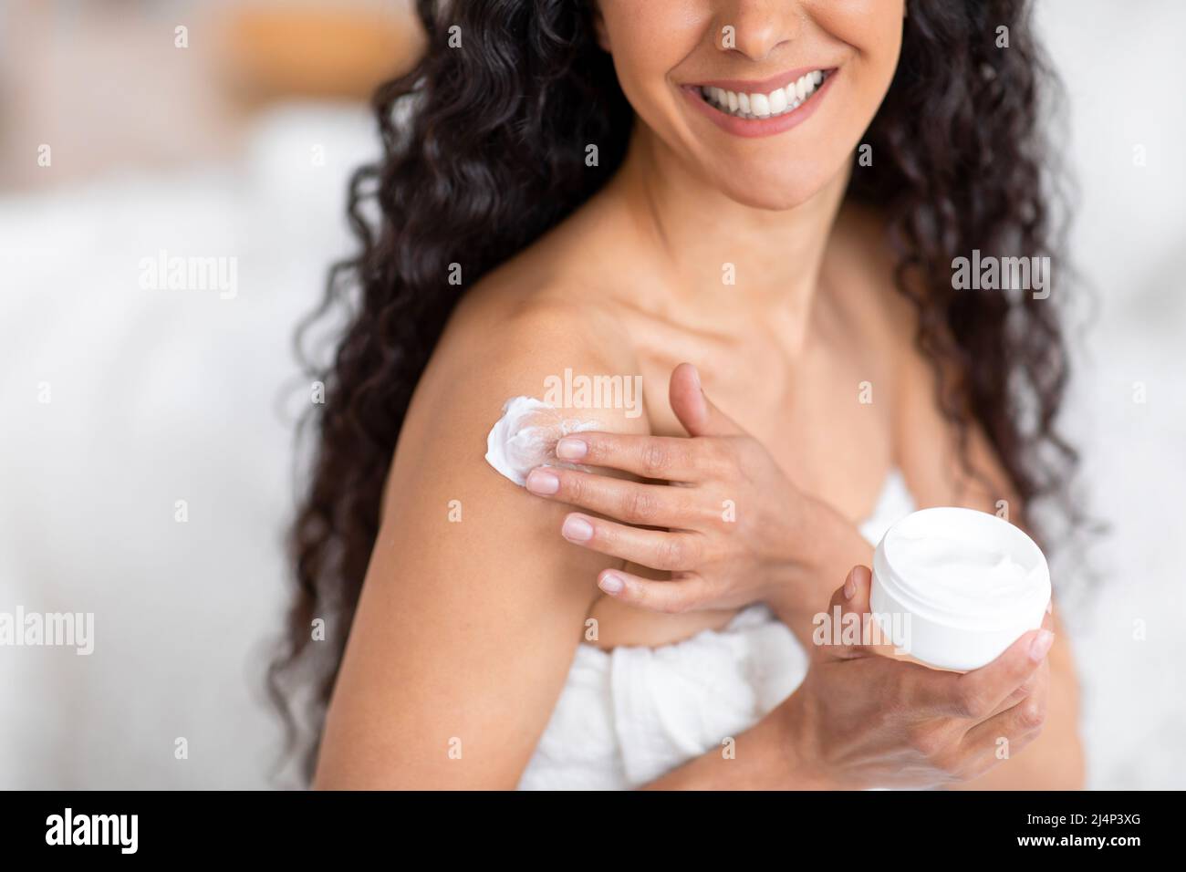Glad young caucasian brunette lady with long curly hair in towel applies cream on shoulder in bedroom interior Stock Photo
