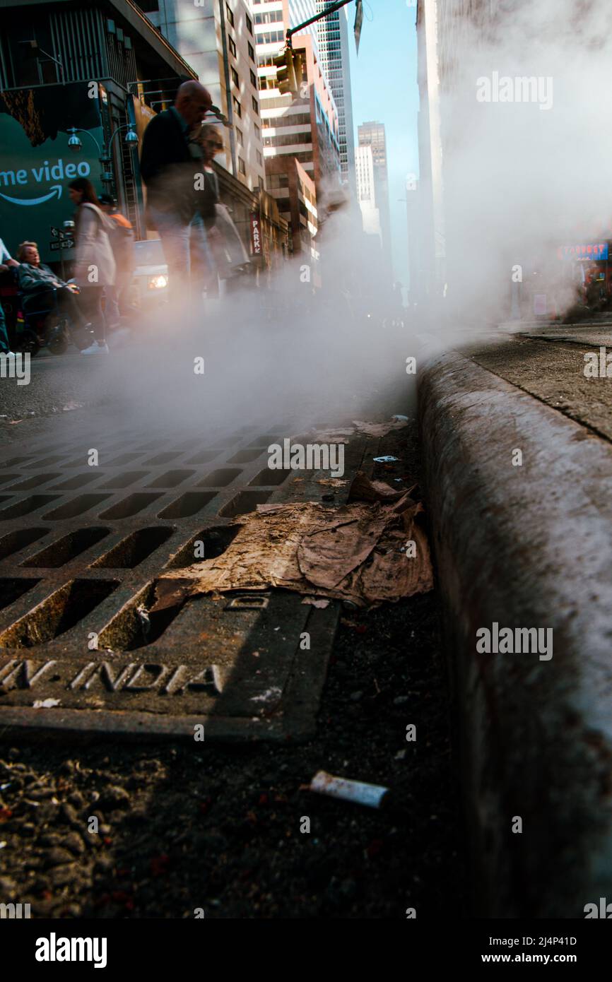 Steam coming out of a sewer grate with cars and people rushing by . Selective Focus Stock Photo