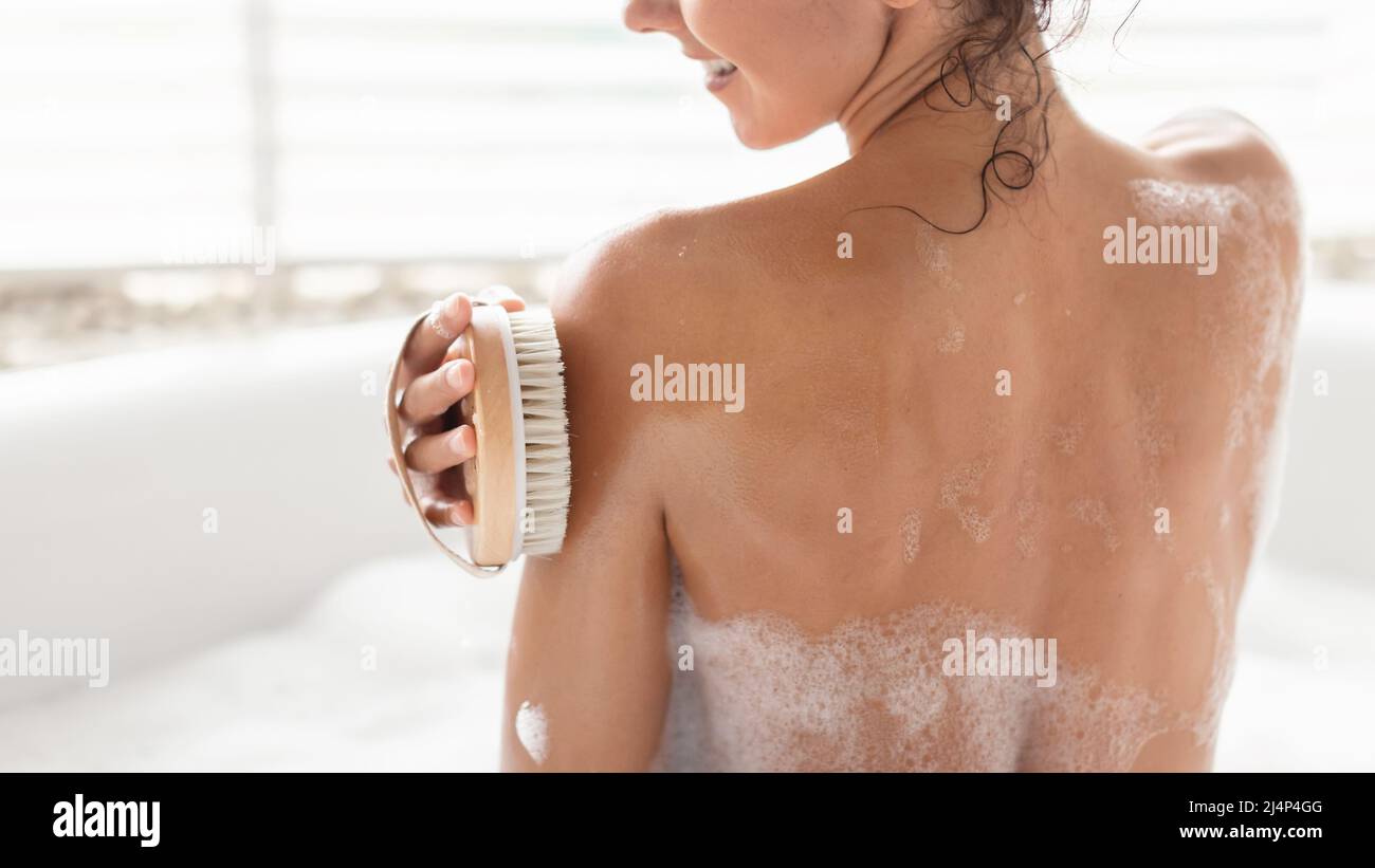 Back view of millennial woman making lymphatic massage with brush, scrubbing skin while taking bubble bath at home Stock Photo