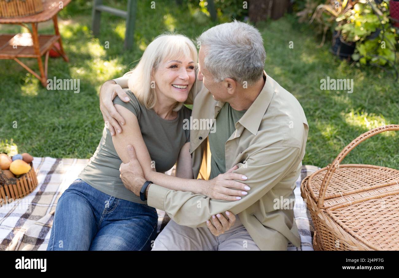 Excited senior spouses having picnic in garden, embracing and laughing, sitting together on blanket outdoors Stock Photo