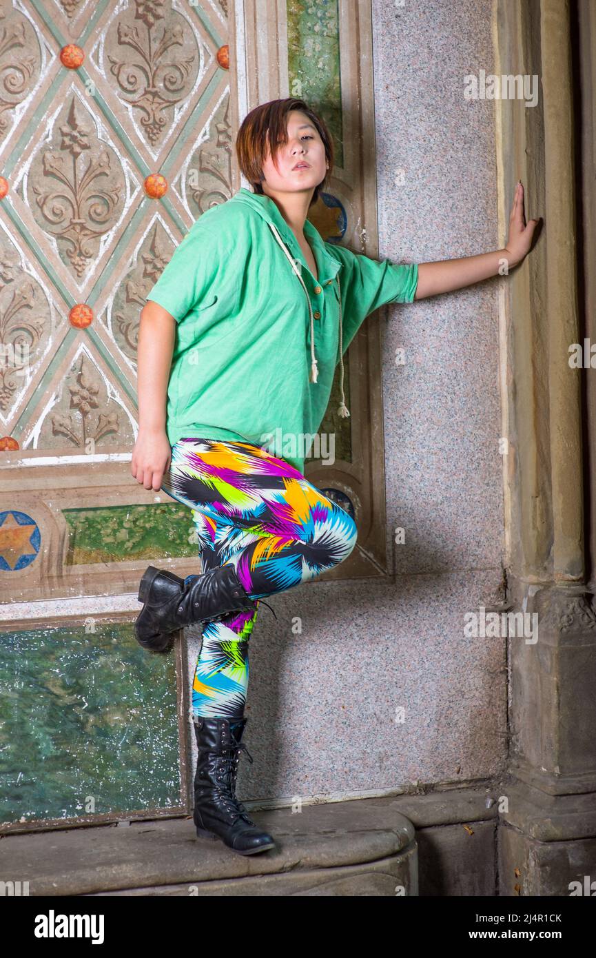 Dressing in a green pull over hoodie,  colorful pants and black leather, long lace boots, a young Chinese girl is standing in the corner of a old styl Stock Photo