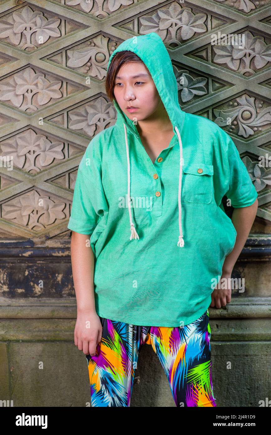 Dressing in a green pull over hoodie,  colorful pants, a young Chinese girl is standing against an old fashion style wall, lost in thought. Stock Photo