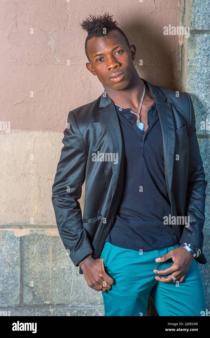Dressing in a black blazer, green pants, a young black guy with mohawk hair is standing against the wall, his head tilted one side, confidently lookin Stock Photo
