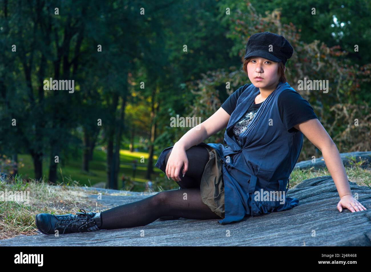 Dressing in a blue sleeveless long blouse, black leggings and boots, wearing a black corduroy cop, a young chinese girl is sitting on rocks, seriously Stock Photo