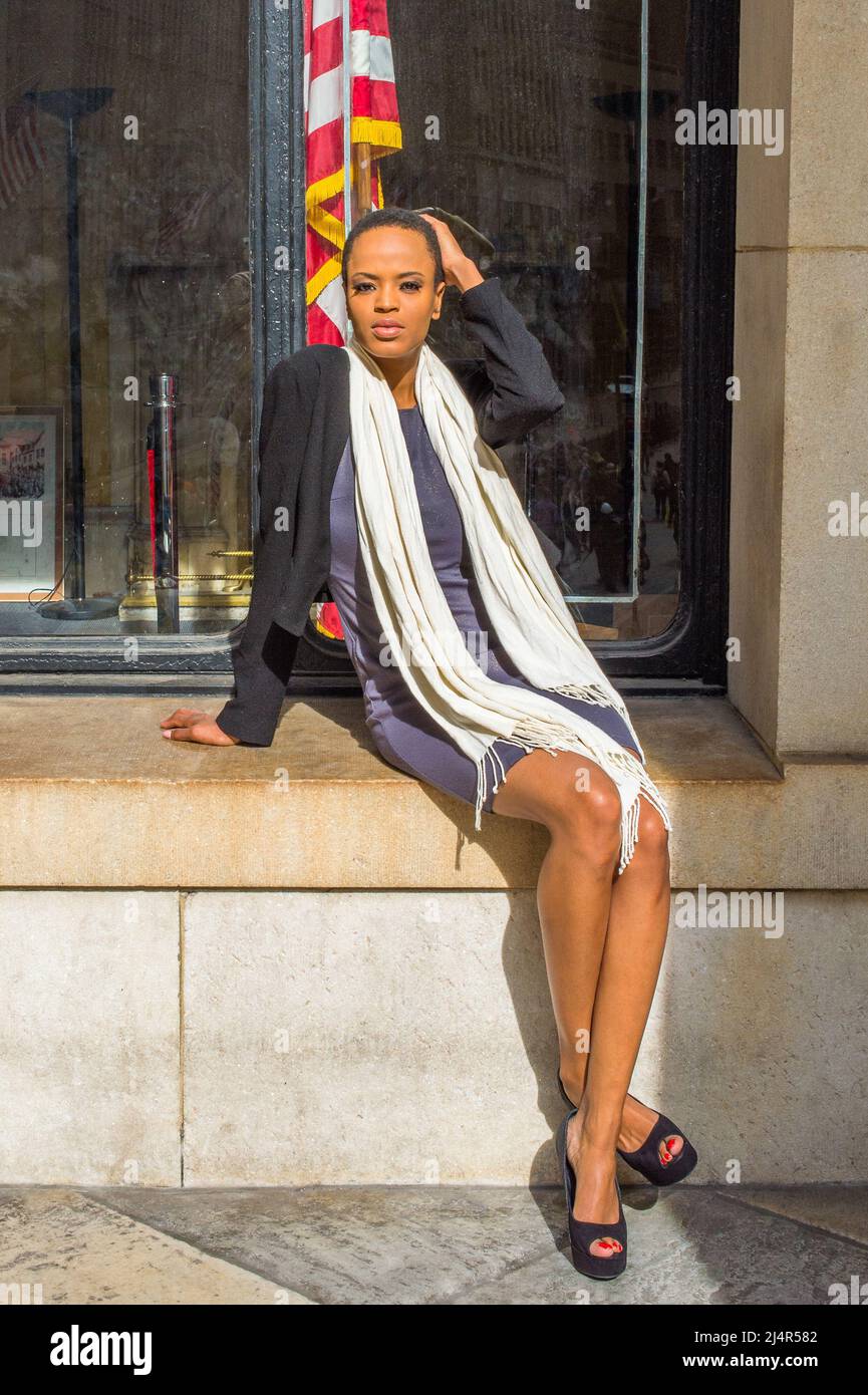 Dressing in a black jacket,  a gray fitted dress, open toe high heels, a long white scarf, a young black businesswoman is sitting on a frame of a wind Stock Photo