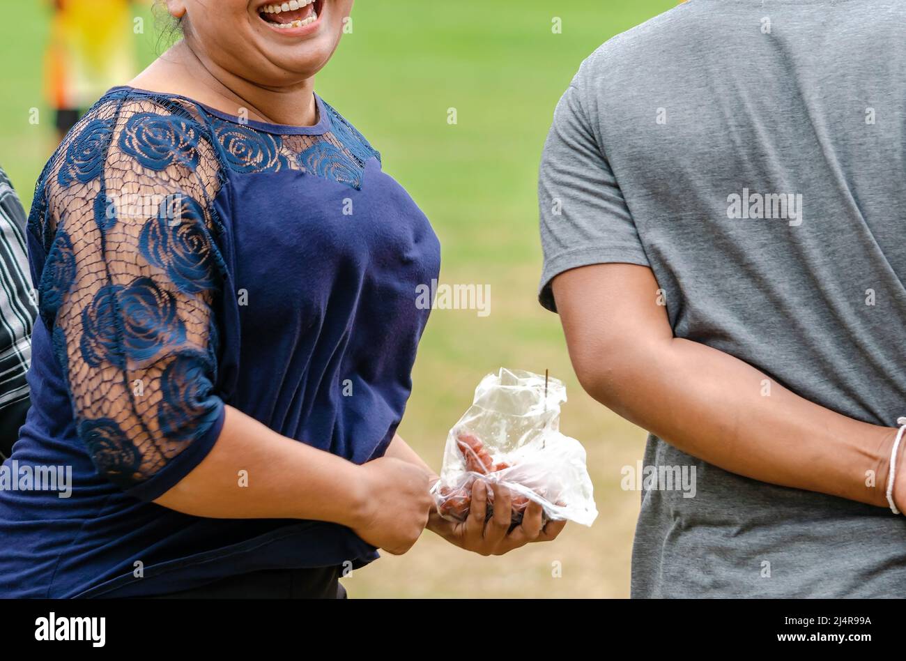 Happiness mature woman in blue dress smiles and laughs during eats sausage in her hand while cheering for sports at outdoor stadium. Thai street food. Stock Photo