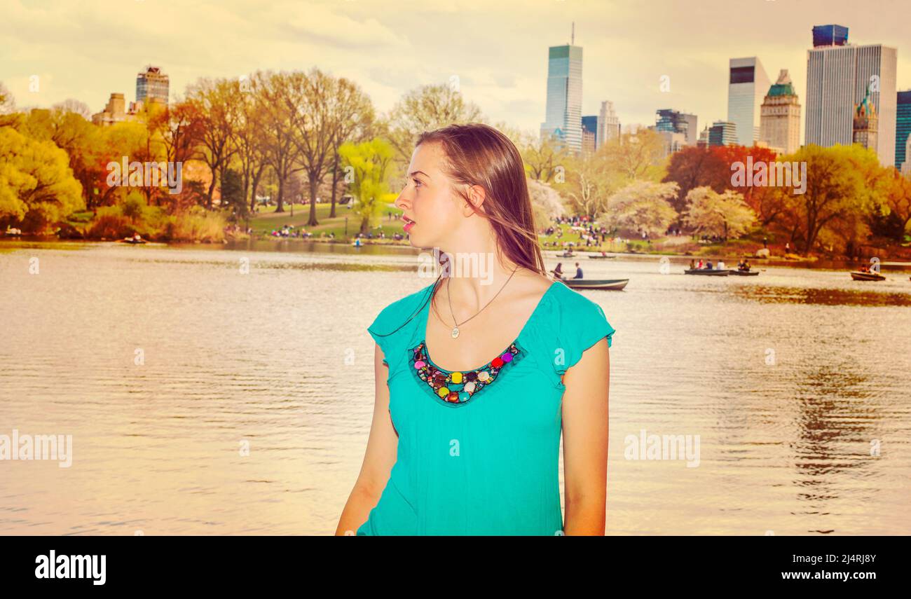 American teenage girl traveling in New York, wearing blue sleeveless clothes, standing by lake at Central Park in spring day, looking around. People r Stock Photo