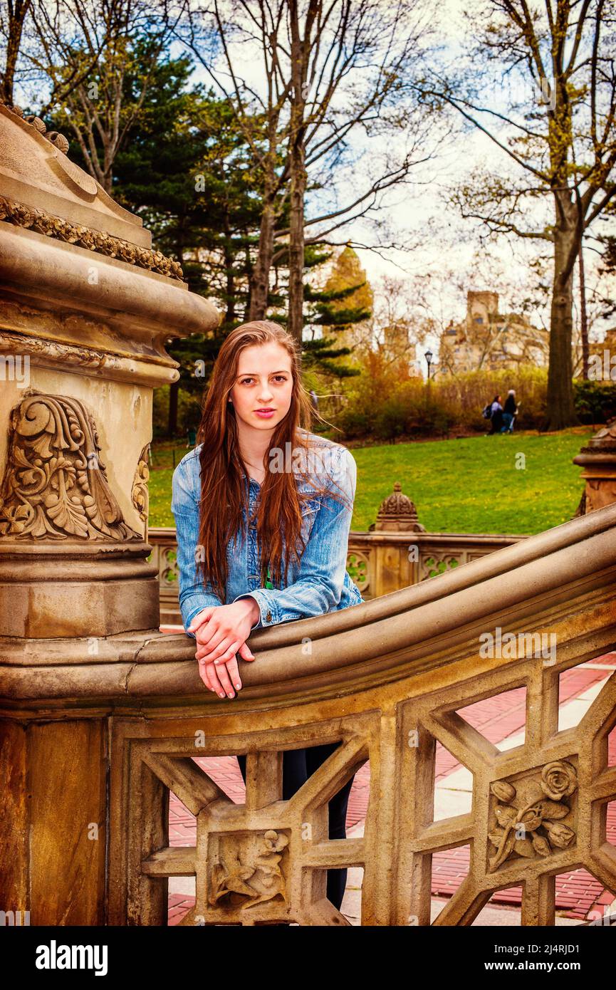 American teenage girl wearing Denim jacket, bending over, arms on old fashion style fence at Central Park, New York, smiling, looking at you. Instagra Stock Photo