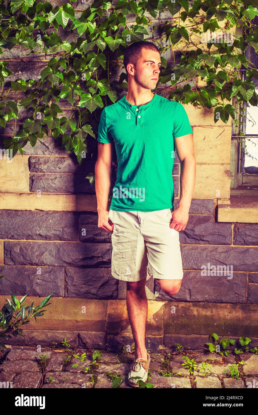 Young man waiting. Wearing a green short sleeve Henley shirt, light yellow shorts, leather casual shoes, bending a leg, a young guy is standing agains Stock Photo