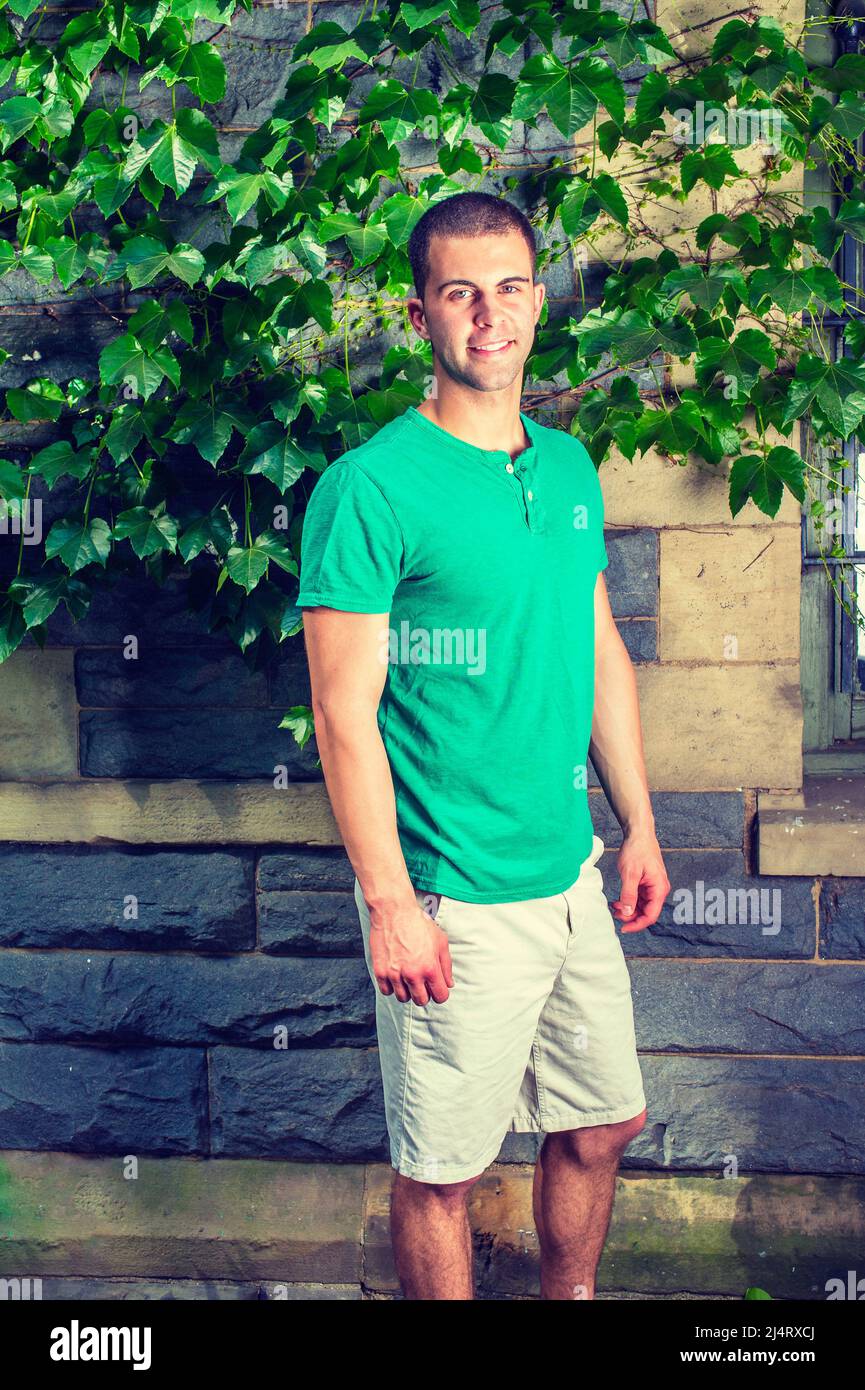 Portrait of young man. Wearing a green short sleeve Henley shirt, light yellow shorts, a young guy is standing by the wall with green ivy leaves, smil Stock Photo