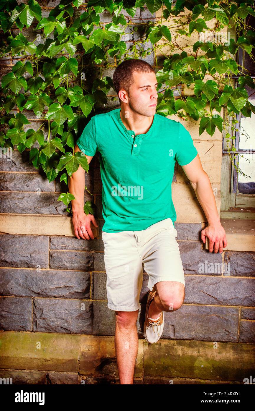 Young man waiting. Wearing a green short sleeve Henley shirt, light yellow shorts, bending a leg, a young guy is standing against the wall with green Stock Photo