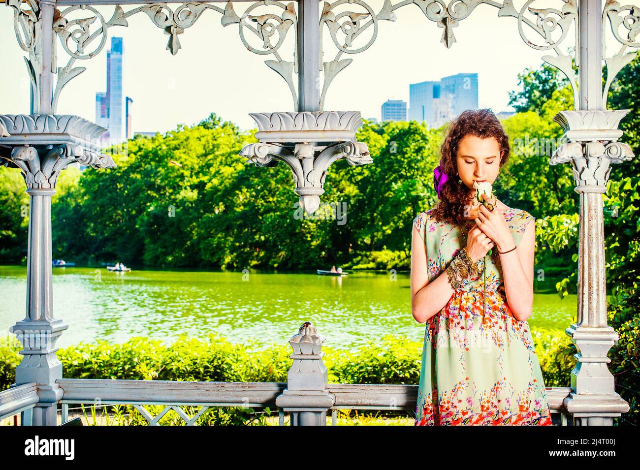 Lady Missing You at Park. Pretty teenage girl wearing sleeveless, long dress, standing inside pavilion, hands holding white rose, looking down, smelli Stock Photo