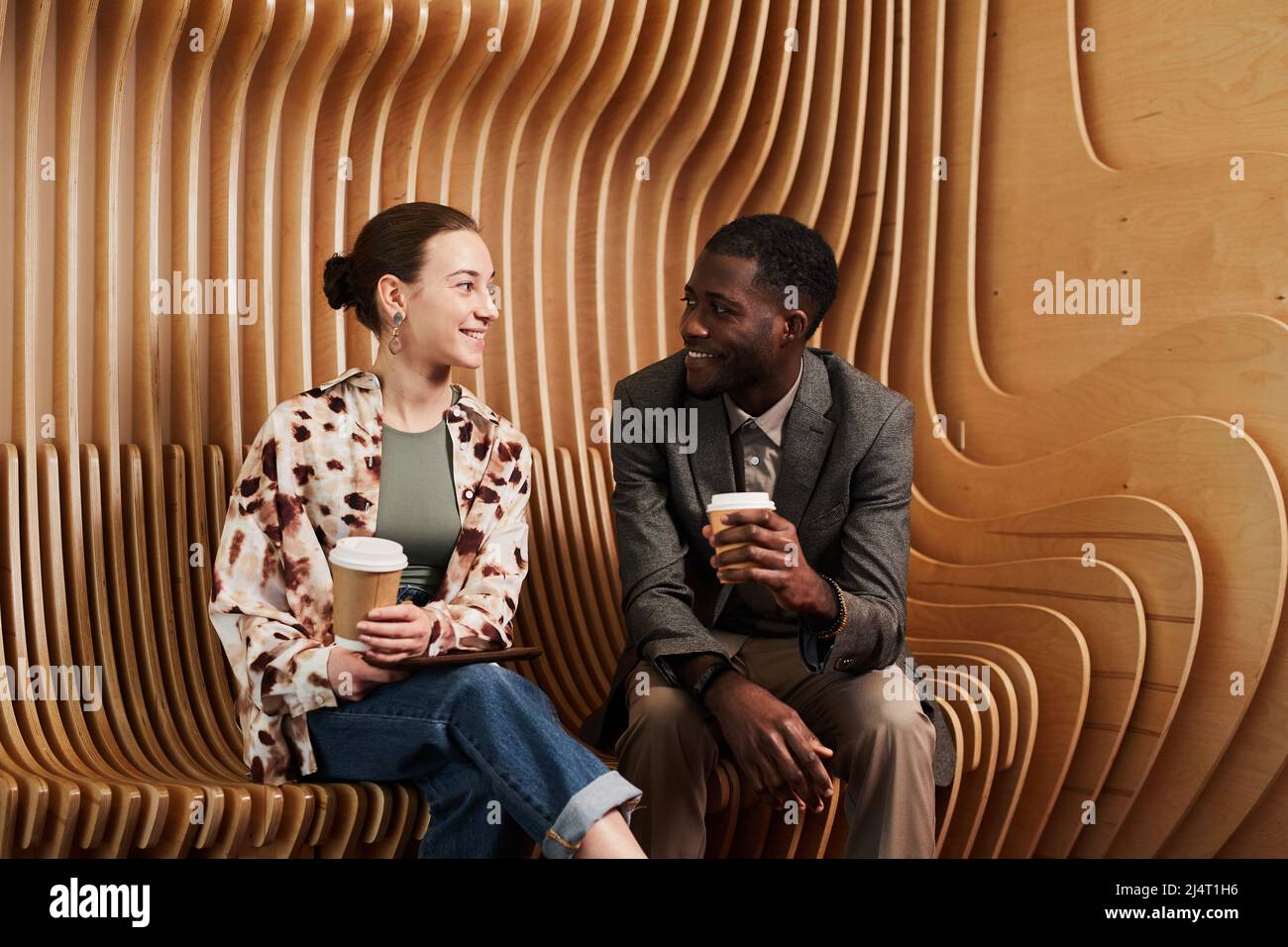 Portrait of to young coworkers chatting while relaxing at lounge in modern office space with wooden panelling Stock Photo