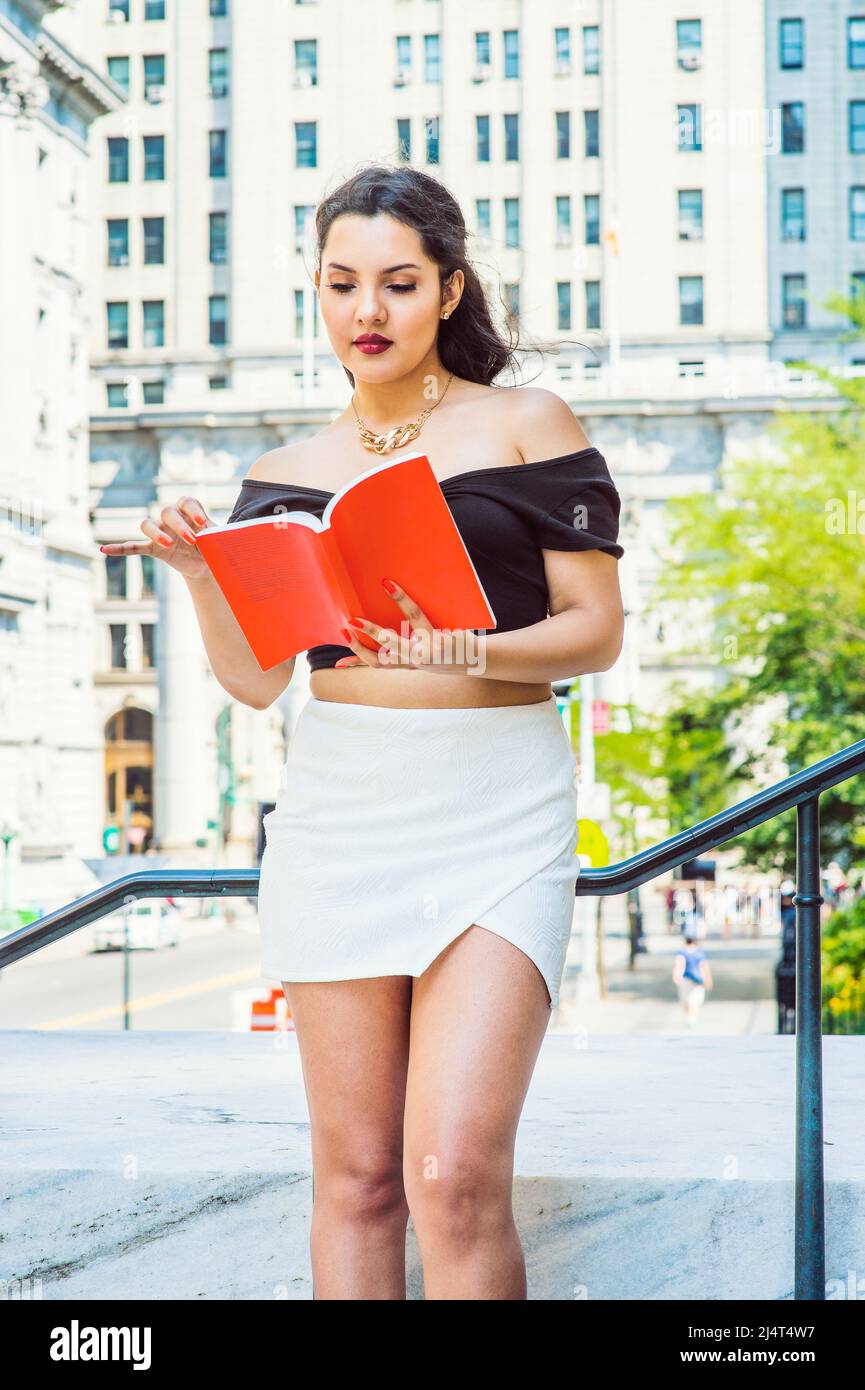 College Student. Dressing in black, short sleeve top, white short wrap skirt, a young pretty girl with long curly hair is standing by railing outside Stock Photo