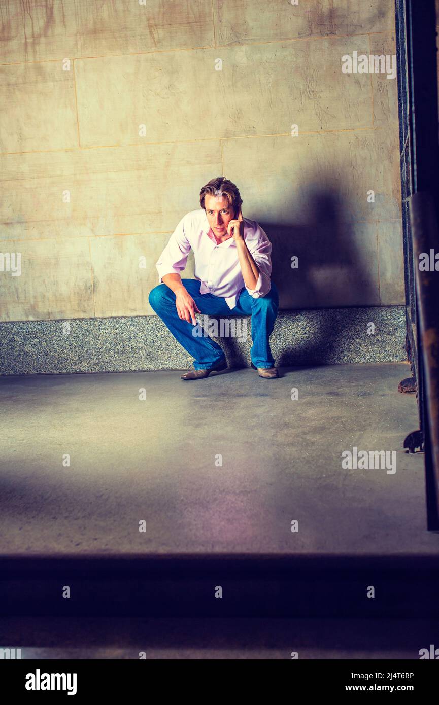 Mad Man. Wearing a light pink, long sleeve shirt, blue jeans, crossing arms, a young man is leaning against a wall in the corner of a metal gate, angr Stock Photo