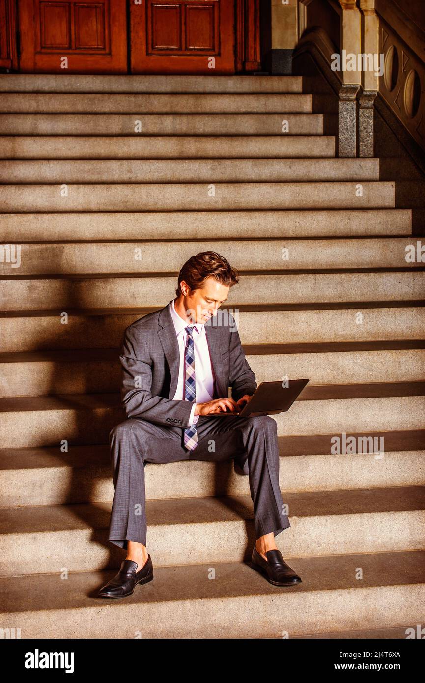 Businessman Working Outside. Dressing formally in gray suit, white under shirt, patterned neck tie, a young businessman is sitting on steps outside an Stock Photo
