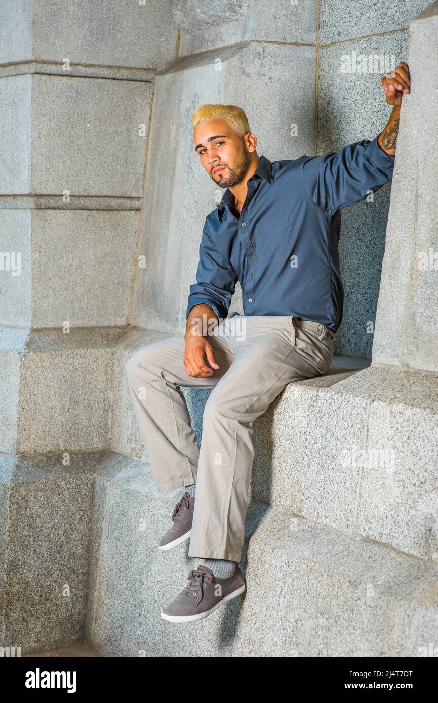 Young Man Relaxing Outside. Wearing a blue shirt, gray pants, casual shoes, a young guy with beard, yellow hair is sitting on a concrete wall, looking Stock Photo