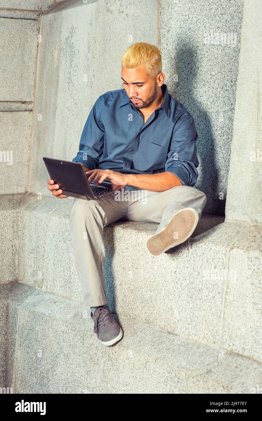 Young Man Working Outside. Wearing a blue shirt, gray pants, casual shoes, a young guy with beard, yellow hair is sitting by a concrete wall, looking Stock Photo