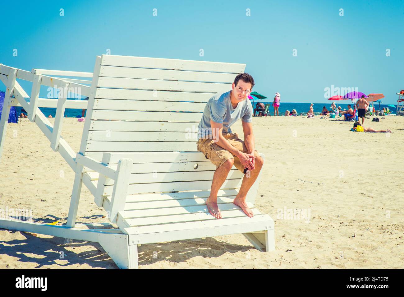 People on the Beach. Wearing a gray t shirt, dark yellow shorts, barefoot, bending back over, holding hands, a young handsome man is sitting on a wood Stock Photo