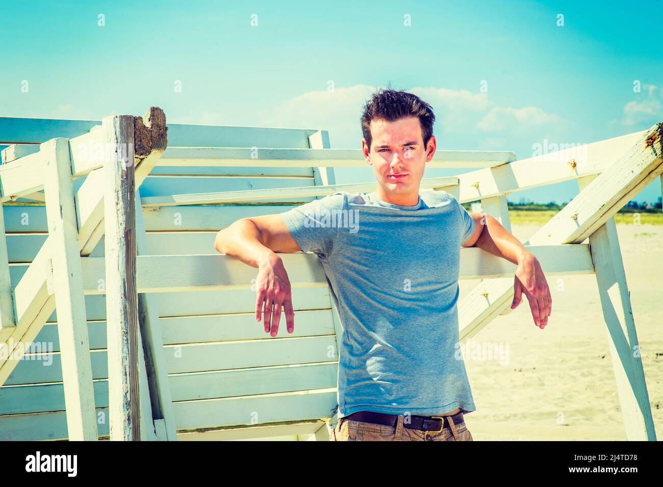 Man waiting for you. Wearing a gray t shirt, arms resting on a wooden stick, a young handsome guy is standing by a wooden structure on the beach, narr Stock Photo