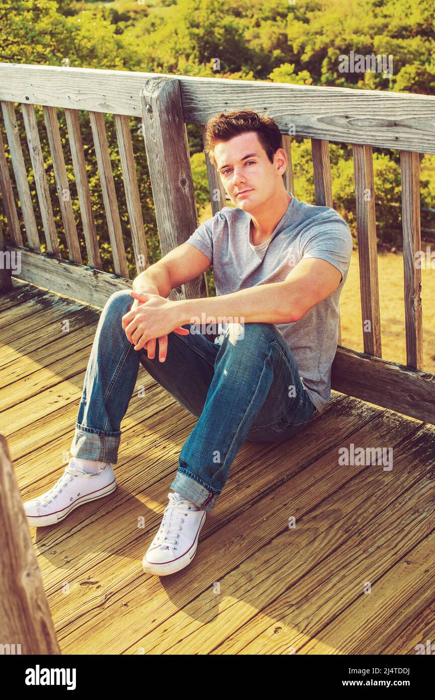 Man Relaxing Outside. Wearing a gray T shirt, jeans, white sneakers, a young handsome guy is sitting on the wooden floor, back against fence in a remo Stock Photo