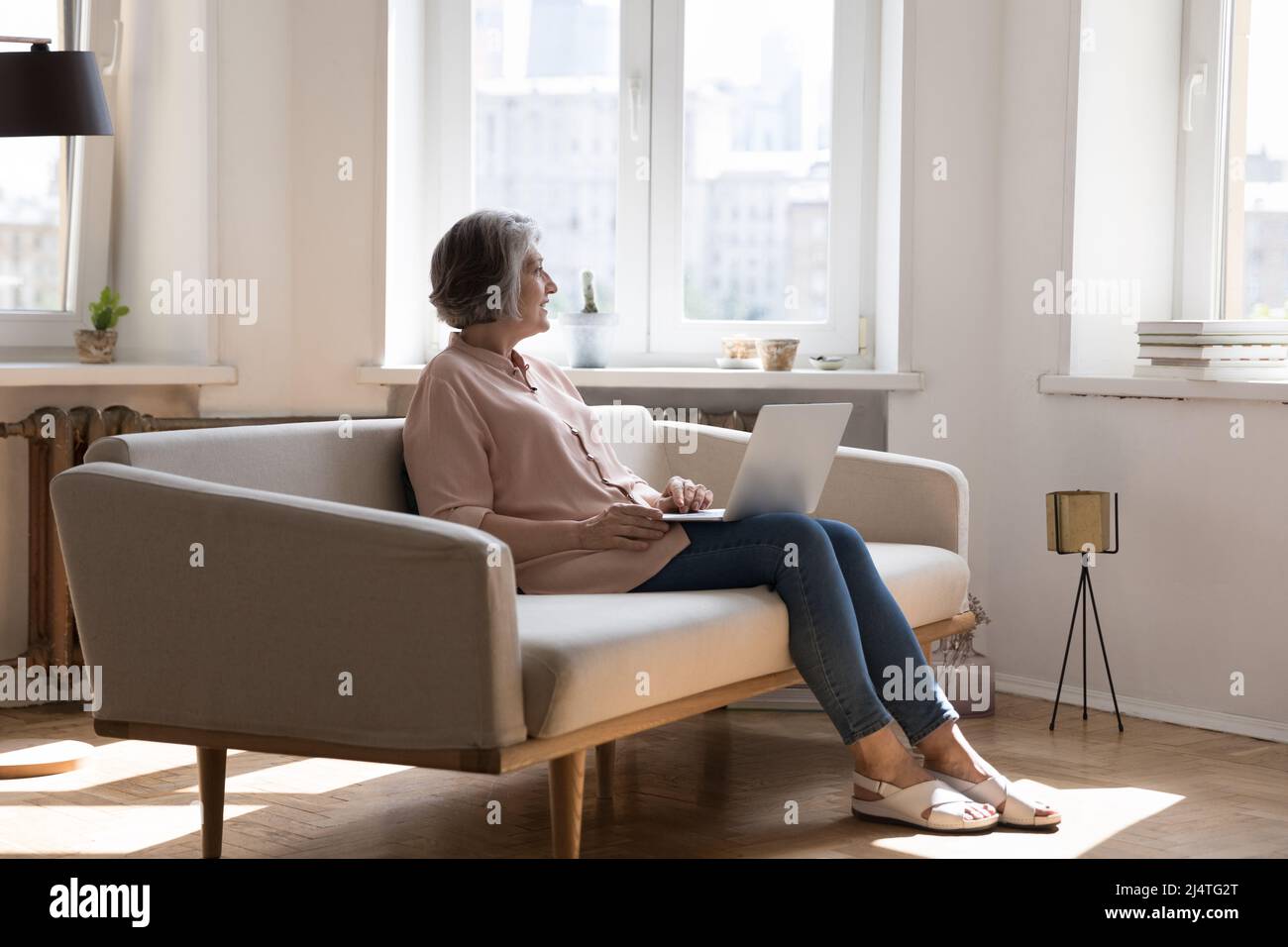 Aged woman sit on sofa with laptop staring out window Stock Photo