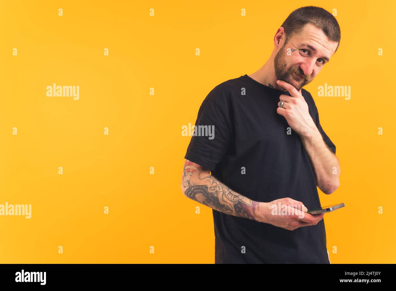 Smiley friendly bearded caucasian guy with tattoos holding smartphone in one hand and touching his beard with the other while looking at camera. Isolated studio shot over yellow background. High quality photo Stock Photo