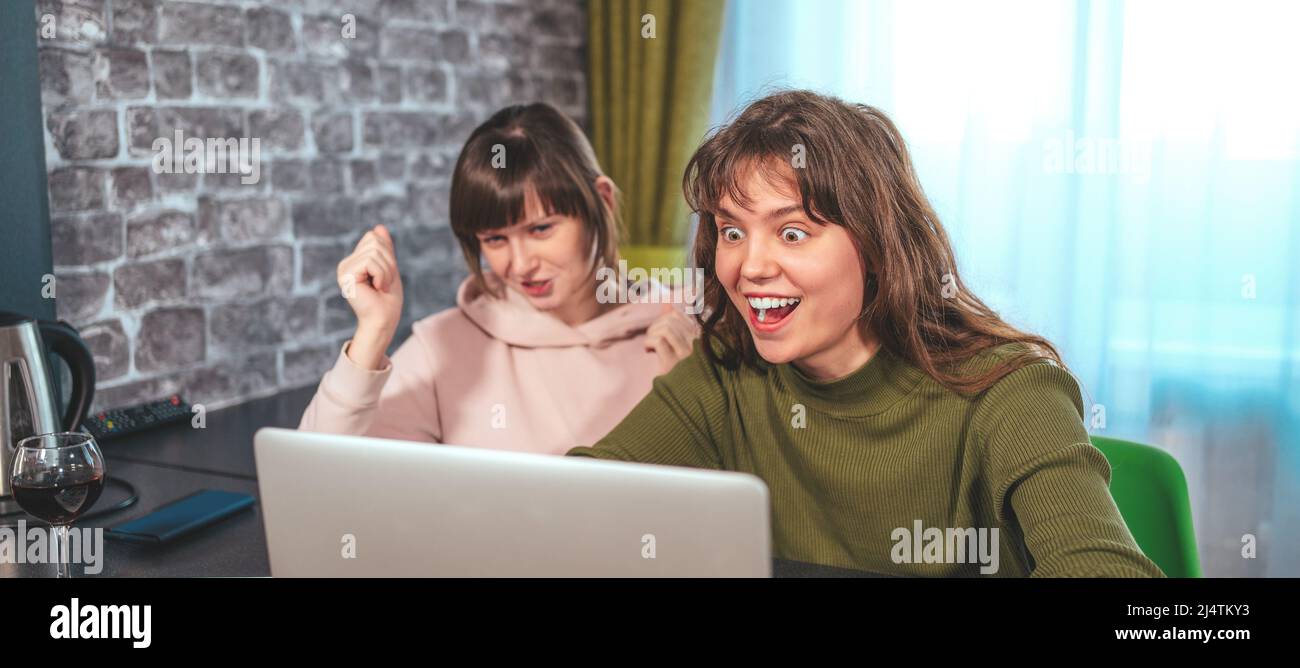 Two girlfriends sitting at laptop begin to rejoice emotionally. Winning championship, celebrating goal scored, winning game concept. Stock Photo