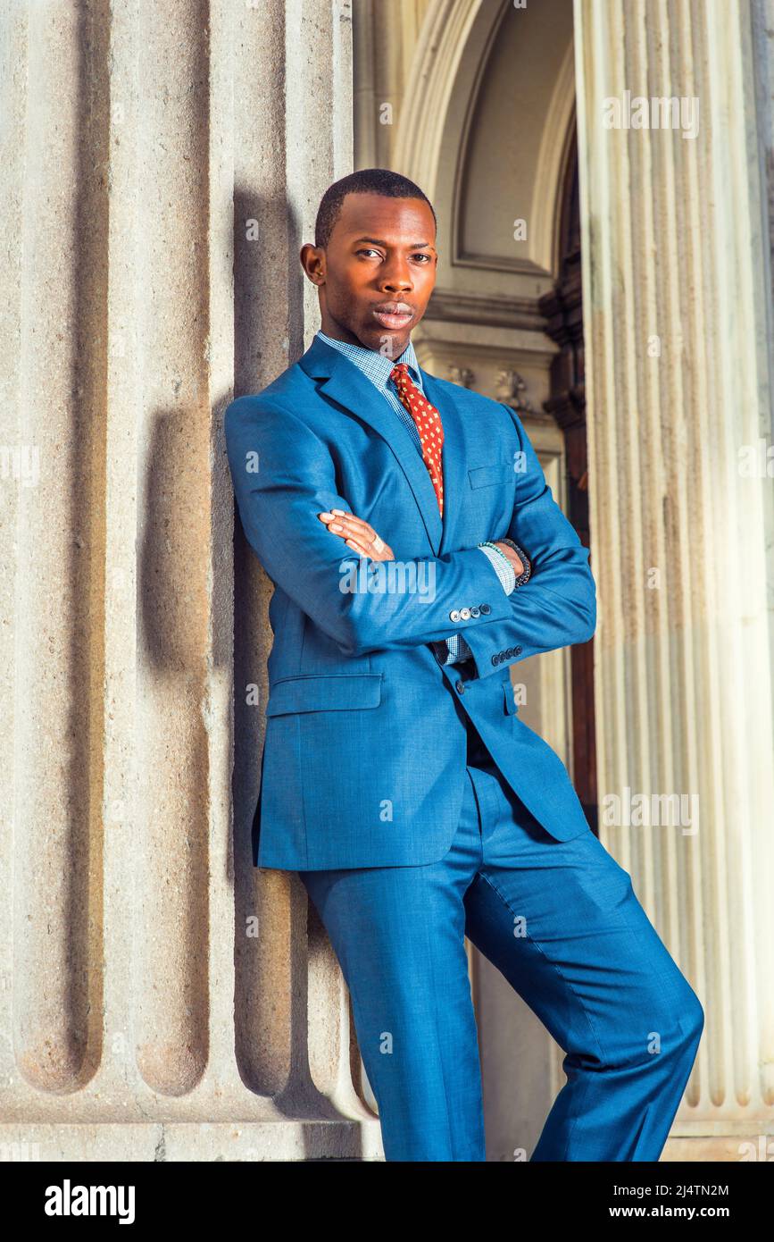 Business Man. Dressing formally in blue suit, patterned undershirt, tie, short haircut, crossing arms, a young black guy is standing by a column outsi Stock Photo