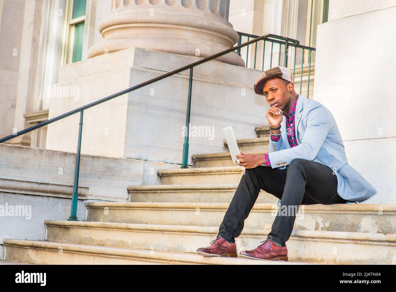 Man Working Outside. Wearing a Apple Newsboy cap, dressing in light gray blazer, black pants, brown leather shoes, a young guy is sitting on stairs ou Stock Photo