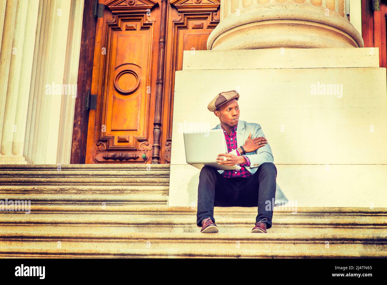 Man Working Outside. Wearing a Newsboy cap, dressing in light gray blazer, black pants, brown leather shoes, a young guy is sitting on stairs outside Stock Photo