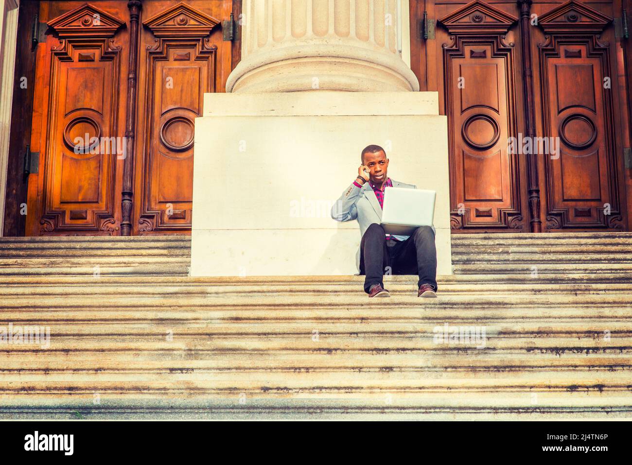 A young black business man is sitting on stairs of old fashion style office building door way, working on laptop computer and talking on his mobile ph Stock Photo
