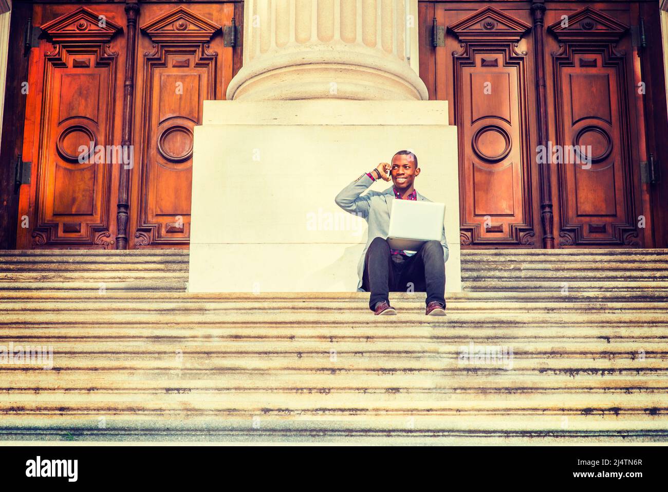 A young black business man is sitting on stairs of old fashion style office building door way, smiling, working on laptop computer, talking on his mob Stock Photo
