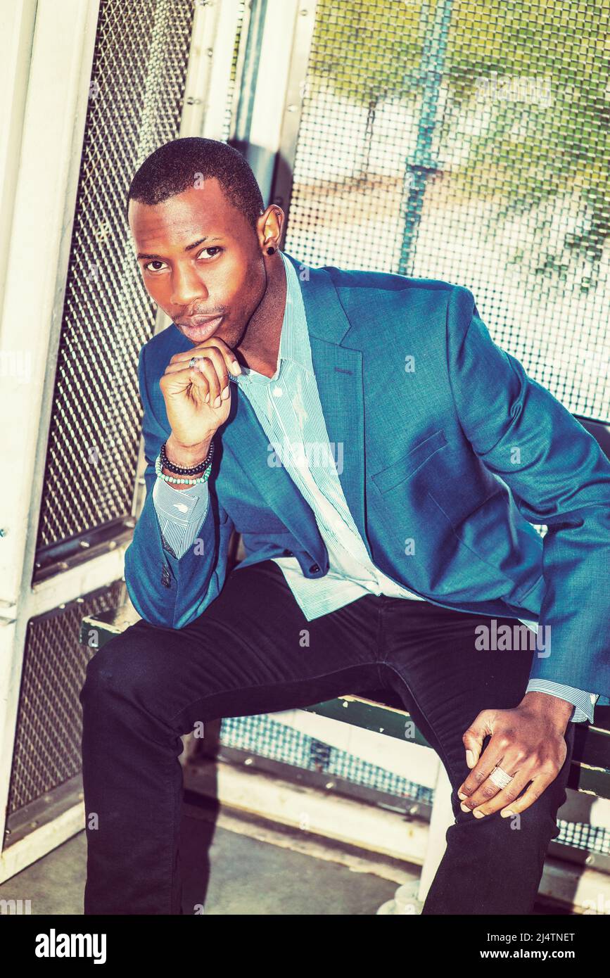 Wearing a blue blazer, black pants,, short haircut, ear stud,  bracelets, finger ring, a young black business man is sitting on bench outside, a hand Stock Photo