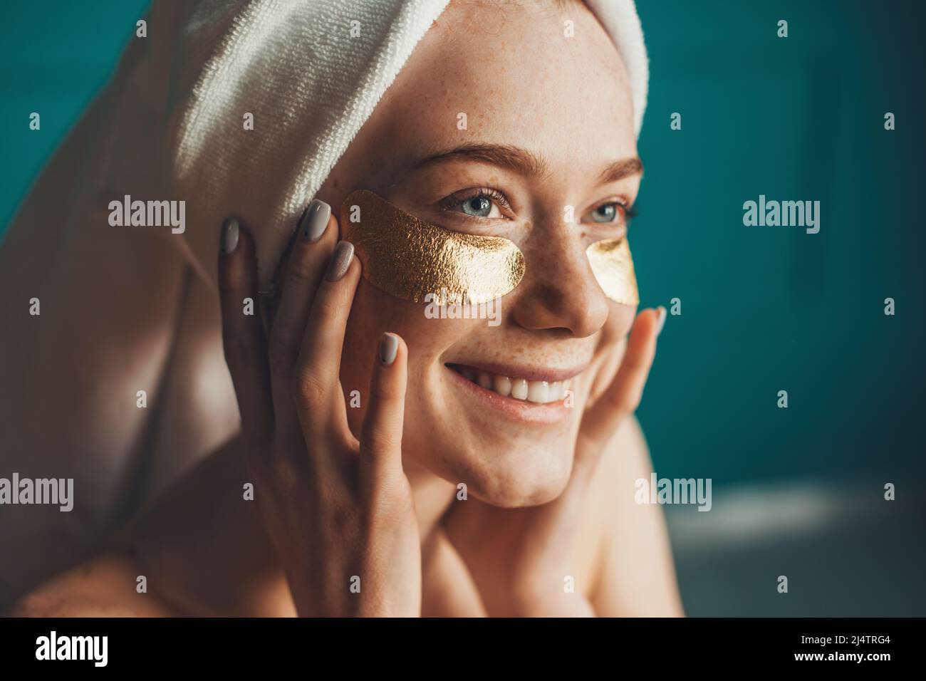 Close-up portrait of a caucasian freckled woman wearing white towel on head, with collagen golden patches under eyes. Girl facial treatment. Facial Stock Photo