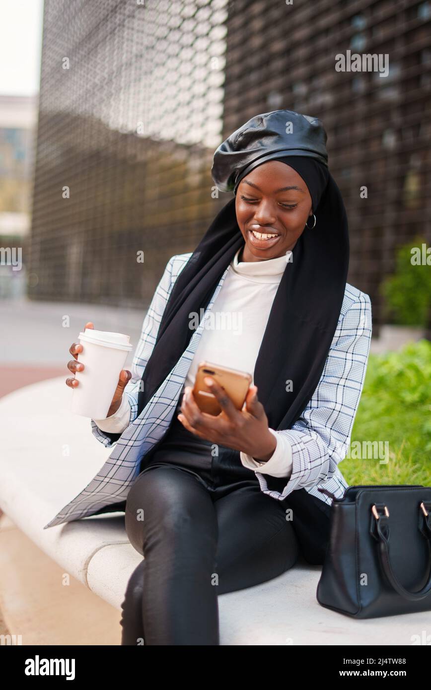 Positive African American female in smart casual clothes and Muslim scarf sitting with crossed legs and having takeaway coffee while watching video on cellphone Stock Photo