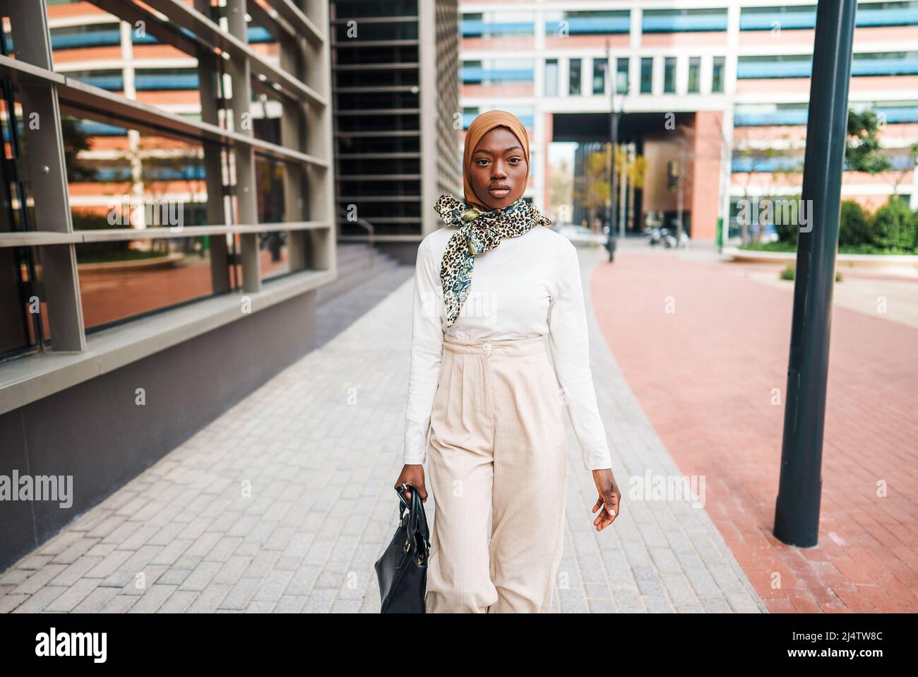 Confident African American female wearing stylish light clothes and hijab walking past modern office towers and looking away Stock Photo