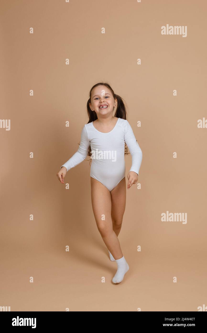 Portrait of young beautiful happy smiling girl with long dark hair in white swimsuit for gymnastics and socks standing on brown background, posing Stock Photo