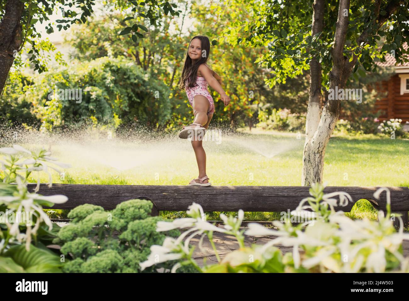 Young cheerful girl walking on log smiling balancing looking at camera with water sprinklers, greenary and house in background in daytime. Dreaming to Stock Photo