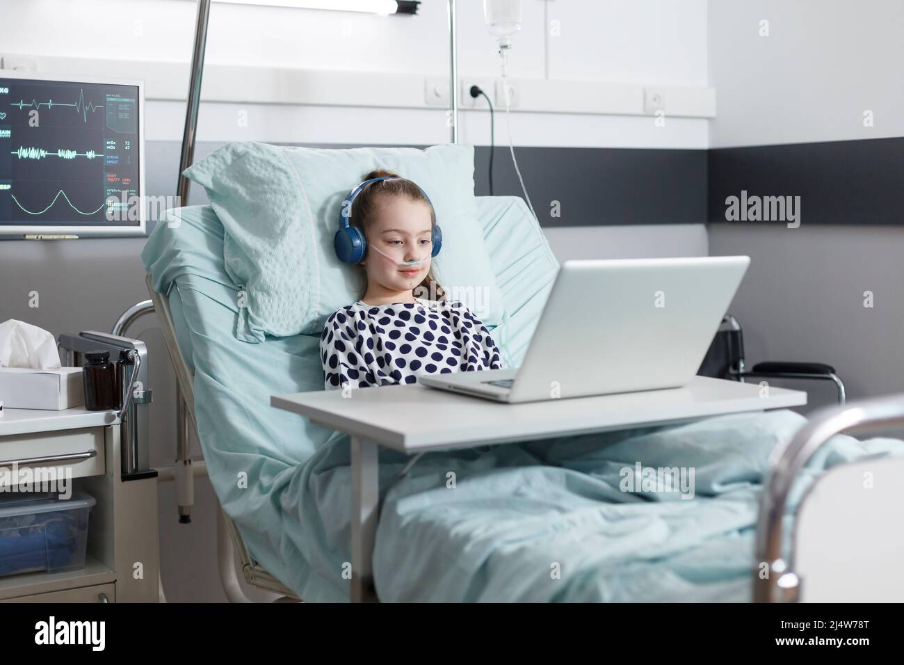 Smiling ill girl patient sitting in pediatric clinic bed while watching amusing cartoons on laptop. Sick little child using computer to watch comical content on internet while sitting in patient ward. Stock Photo