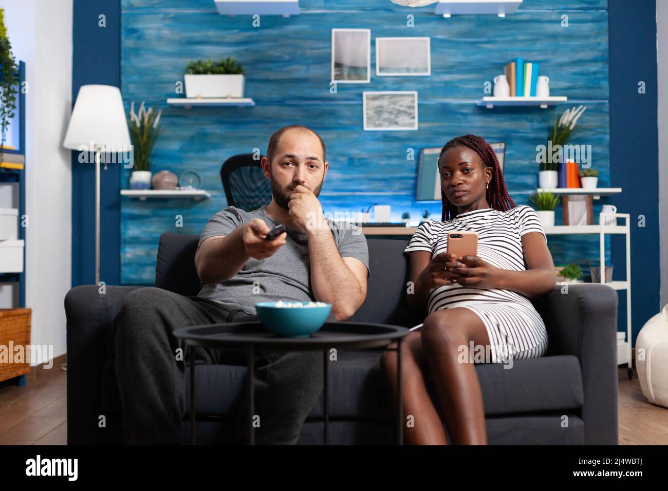 Relaxed future father switching television channels using remote and enjoying popcorn while pregnant woman sitting on sofa and holding smartphone in hand. Diverse couple sitting on couch at home and watching movies on TV Stock Photo